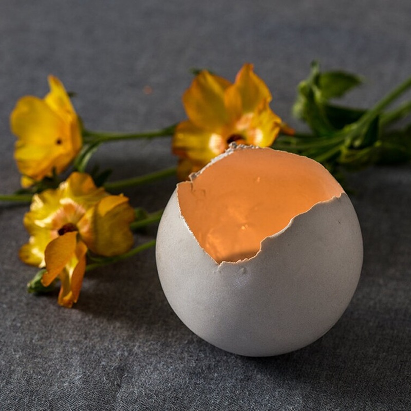 Lit Ceramic Eggshell-Shaped Tealight Holder with Yellow Flowers in Background